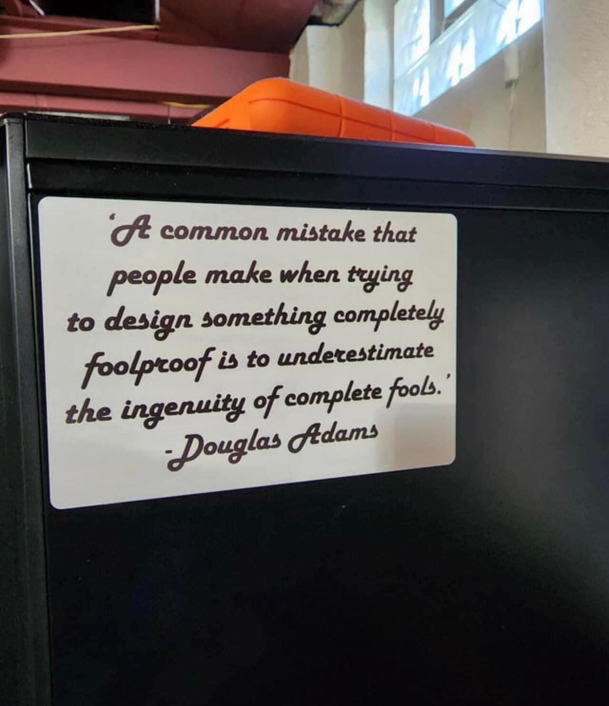 Sign on side of desk quoting Douglas Adams: "A common mistake that people make when trying to design something completely foolproof is to underestimate the ingenuity of complete fools."