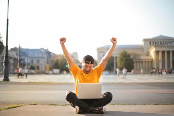 man in yellow crew neck t-shirt and black pants sitting on sidewalk happy with both hands in the air